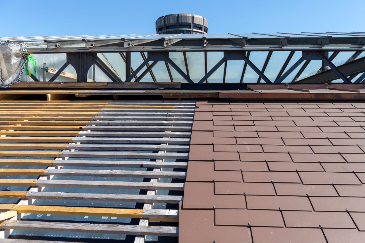 Das Foto zeigt die Erneuerung der Dacheindeckung der Perronhallen, beziehungsweise links das ungedeckte Dach, rechts das Dach mit den neuen Eternitschindeln.