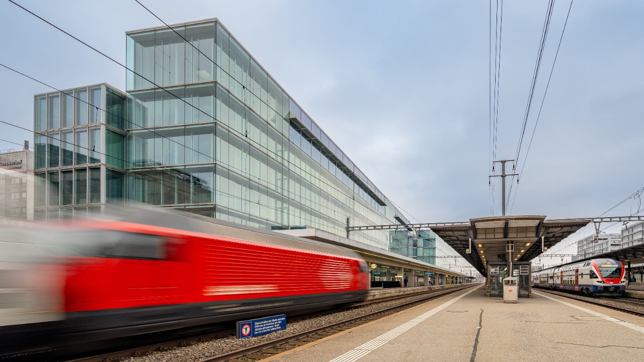 Eine rote Schnellzugslok fährt an einem Bahnhof vorbei, auf der Gegenseite wartet ein Regionalzug. 