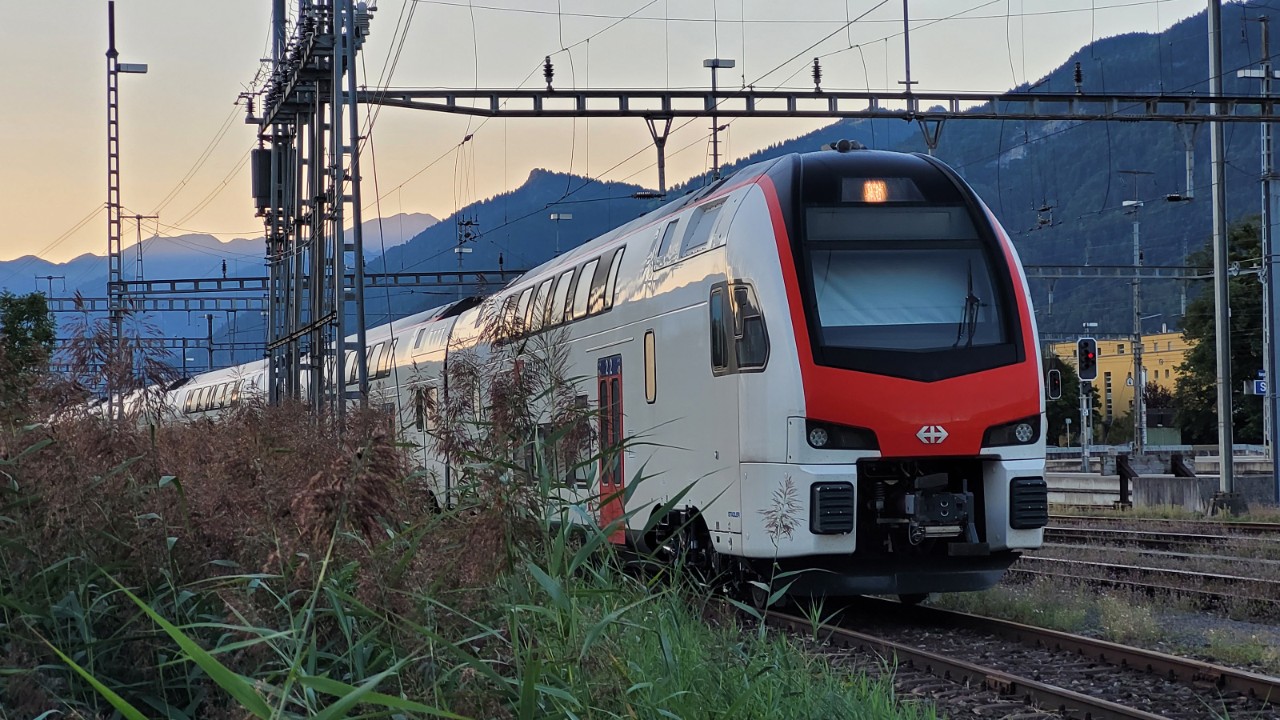 Ein IR-Dosto-Zug steht abgestellt in einem Gleisfeld, im Hintergrund geht die Sonne hinter den Bergen unter. 