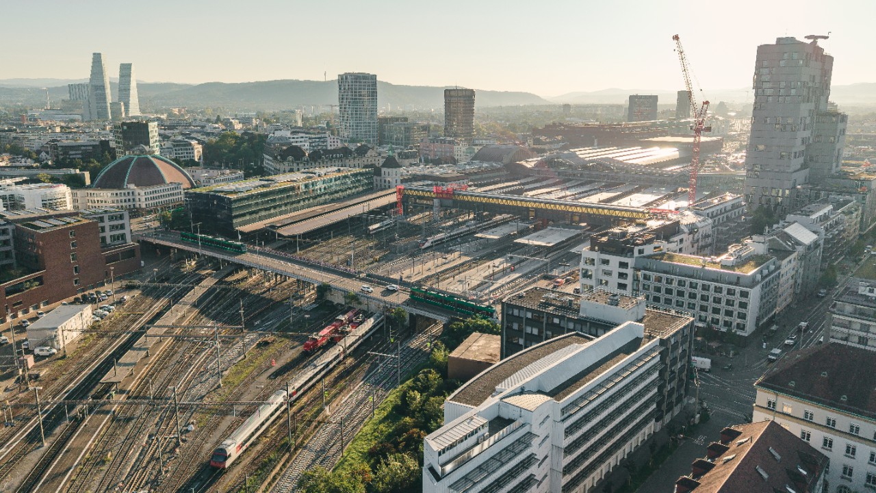 Luftaufnahme des Bahnhofs Basel SBB mit zahlreichen Gleisen, Perrons und abgestellten sowie fahrenden Zügen. Brücken, Betriebsanlagen und Baustellen mit Kränen durchziehen das Areal, umgeben von dicht bebautem Stadtgebiet. Im Hintergrund sind Hochhäuser, weitere Stadtquartiere und Hügelzüge erkennbar.