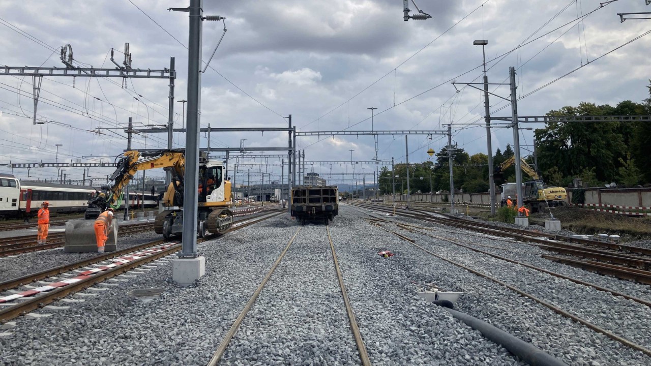 Auf dem Bild sind Arbeiter in orangefarbenen Sicherheitswesten und Helmen zu sehen, die auf einem mit Schotter bedeckten Bahngleis arbeiten. Sie sind von Gleisen, Oberleitungen und einem Bagger umgeben.