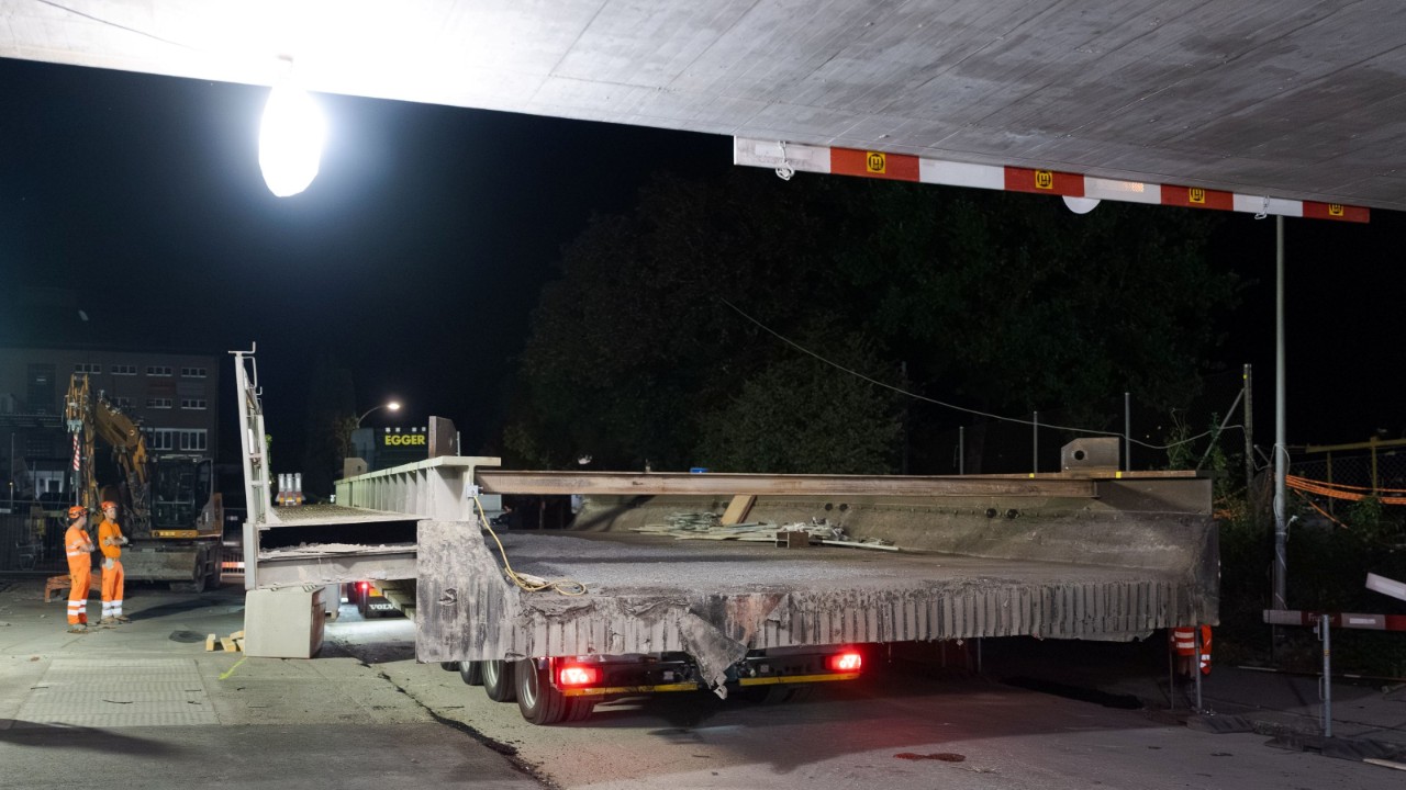 Blick von hinten auf einen grossen Betonbrückenträger, der auf einem Tieflader transportiert wird; am linken Bildrand stehen zwei Arbeiter in Warnkleidung. Die Baustelle ist nachts beleuchtet, im Vordergrund ist die abgesenkte Fahrbahn sichtbar.