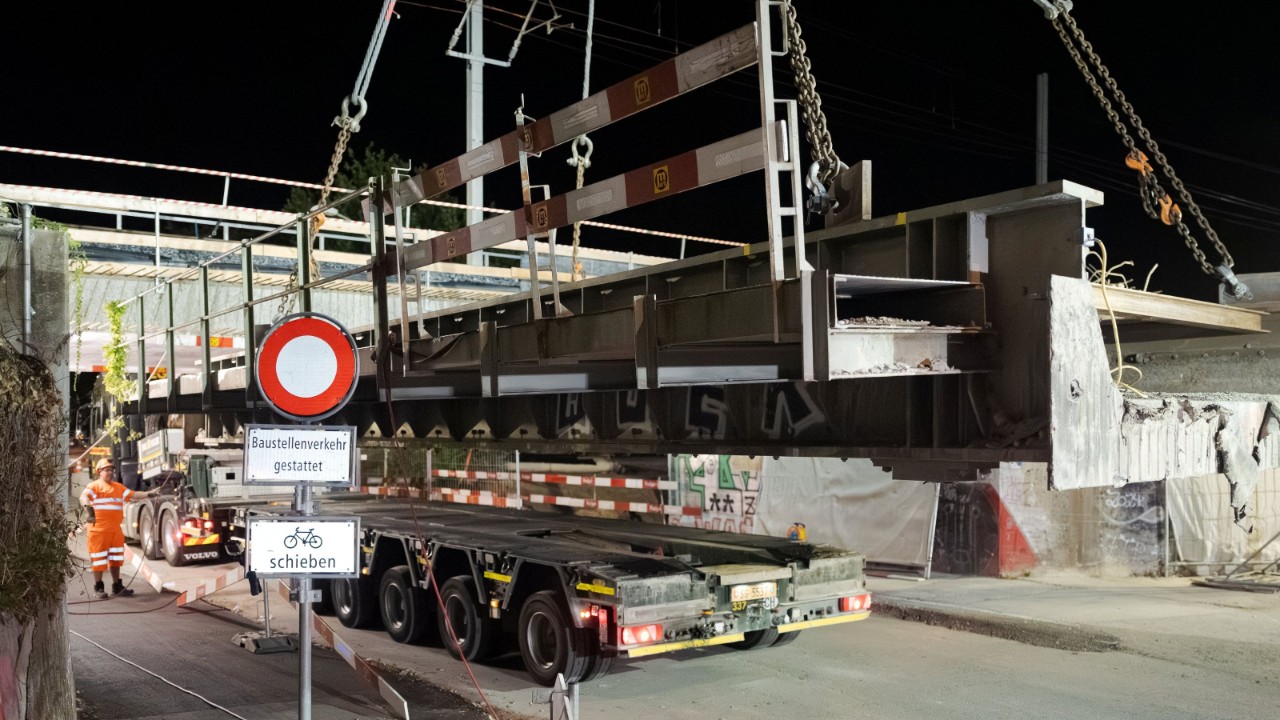Seitenansicht eines schweren Stahl- oder Betonträgers, der mit Ketten am Kran hängt und gerade über einen Tieflader abgesetzt wird. Ein Verkehrsschild und Absperrungen markieren die Baustelle; im Hintergrund stehen Arbeiter in Warnkleidung.