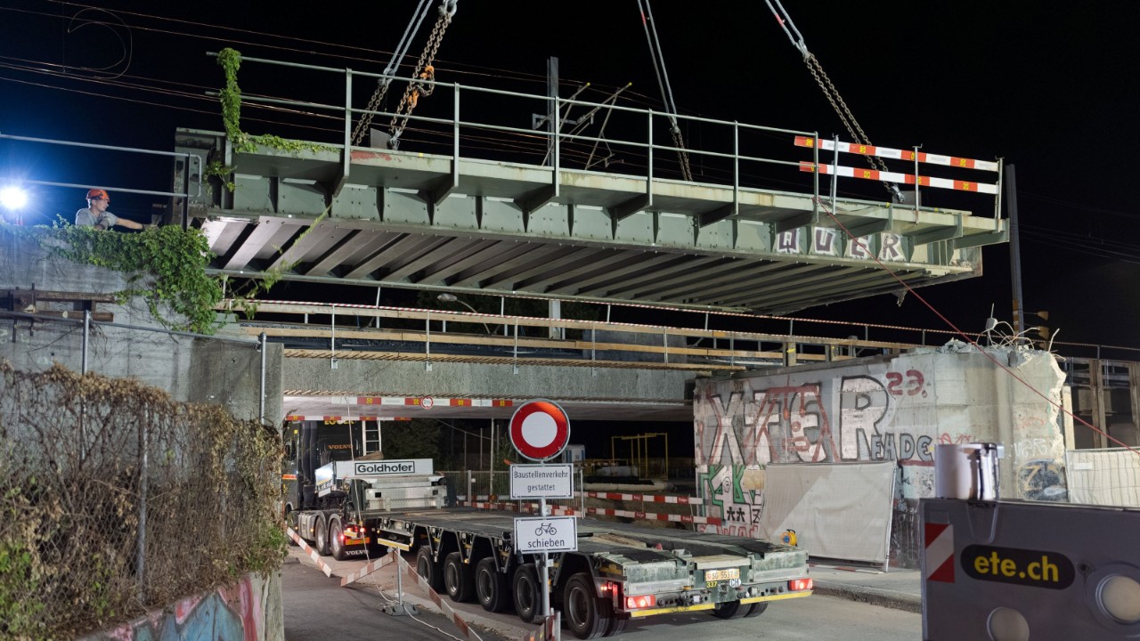 Detailaufnahme des abgesenkten Brückenträgers, der am Kran hängt; zwei Hebeseile sind sichtbar. Im Vordergrund ein Teil des Tiefladers und Baustellenmaterial, im Hintergrund Absperrungen und Graffiti an den Brückenwänden.