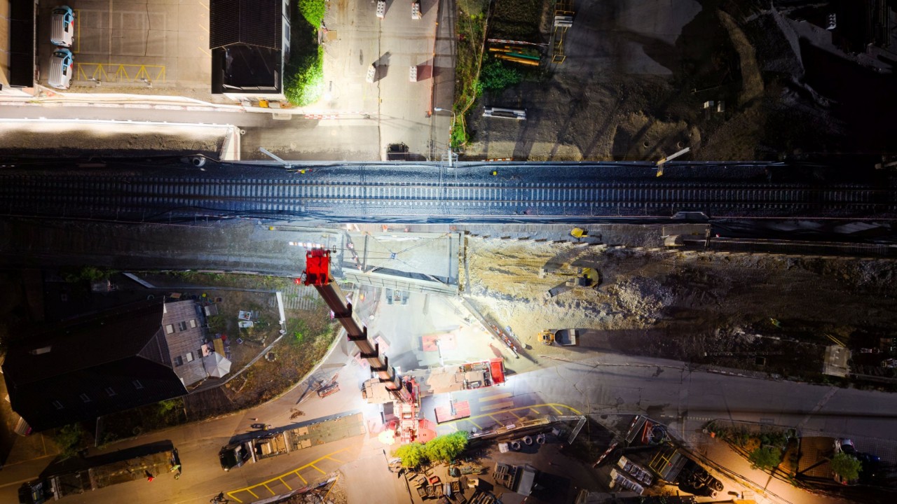 Weitwinkel-Nachtaufnahme der Baustelle Stöckacker: Ein grosser Mobilkran hebt ein Brückenteil über der gesperrten Strasse nahe den Bahngleisen. Baustellenbeleuchtung, Baufahrzeuge und vereinzelte Arbeiter sind um den Kran herum sichtbar; im Hintergrund zeigt sich die Stadt bei Nacht.