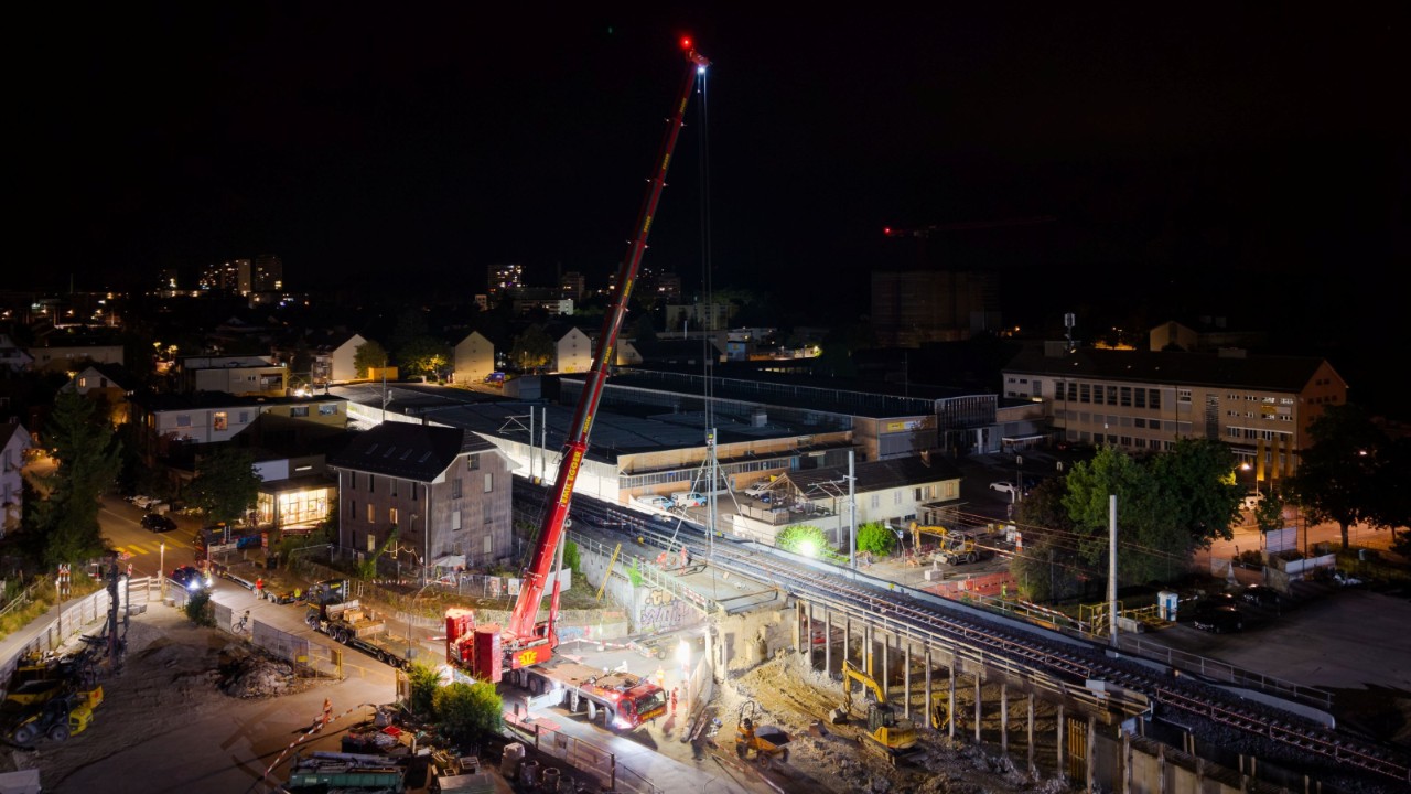 Drohnenaufnahme von oben: Der Mobilkran steht unter dem Bahntrassee und hebt ein Brückenelement, das quer über die Strasse liegt. Gut erkennbar sind die Gleise, Baustellenfahrzeuge, Materiallager und die beleuchtete Arbeitszone in der Nacht.