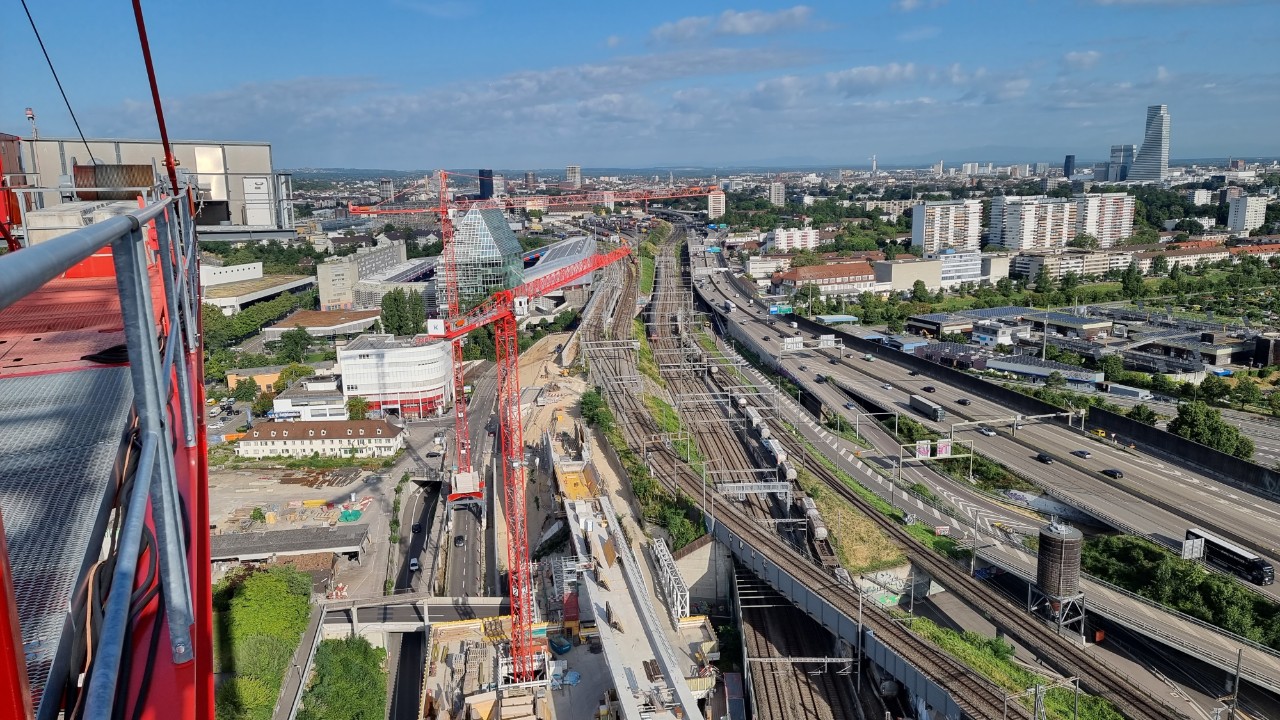 Das Bild zeigt eine Panorama-Sicht von Kran 3 in Muttenz in Richtung Basel, markant sichtbar ist rechts im Bild der Roche-Turm. Im Vordergrund sind die Kräne 2 und 1 unseres Bauprojekts sowie die Baustelle der Donnerbaumbrücke sichtbar.