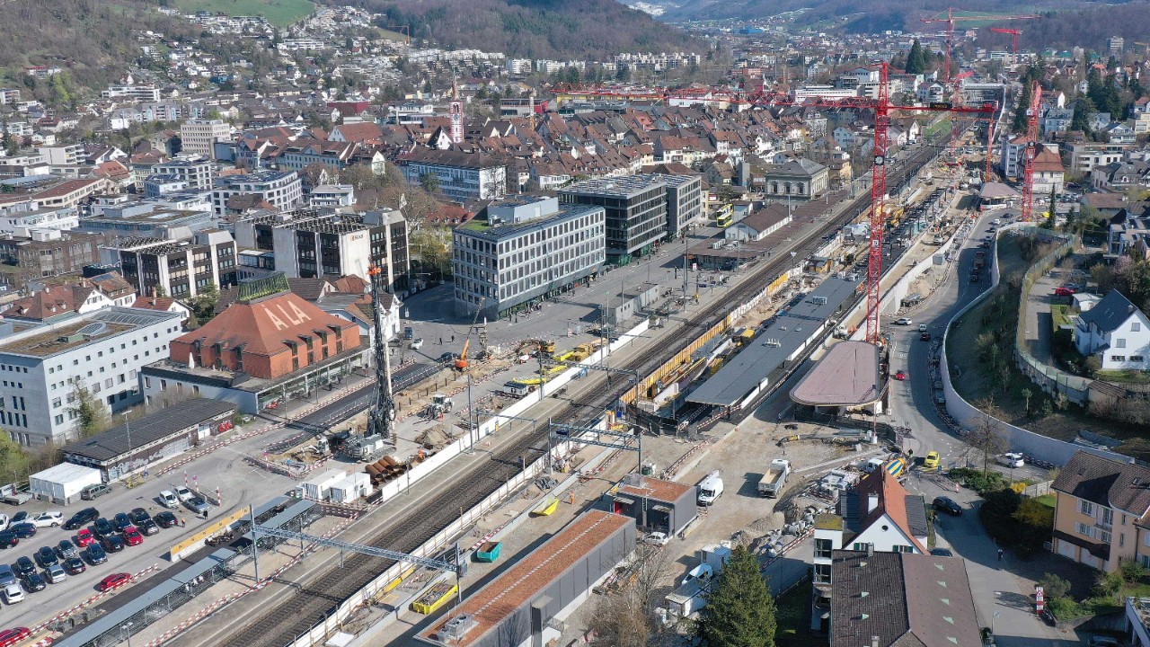 Luftaufnahme eines großen Bahnhofsbereichs in einer Stadt mit umfangreichen Bauarbeiten. Mehrere Gleise und Perrons sind sichtbar, umgeben von Kränen, Baustellenfahrzeugen und Gebäuden. Im Hintergrund erstreckt sich ein dicht bebautes Stadtgebiet mit Hügeln.