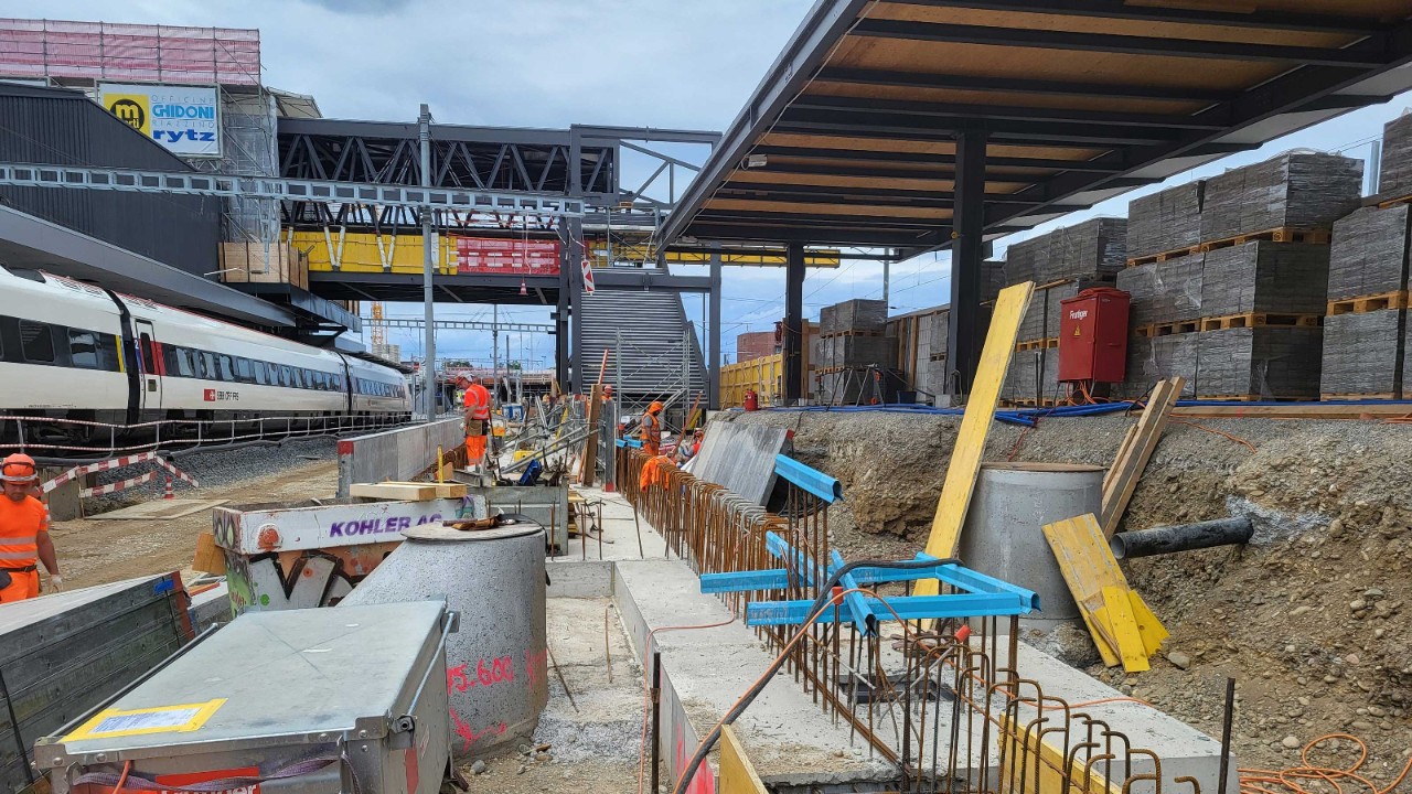Baustelle an einem Bahnhof mit sichtbaren Baugruben, Betonfundamenten und Baumaterialien. Mehrere Bauarbeiter arbeiten neben einem abgestellten Zug, im Hintergrund sind Perrons und eine Passerelle zu sehen. Stapel von Baumaterialien und Baugeräte prägen den Bereich.