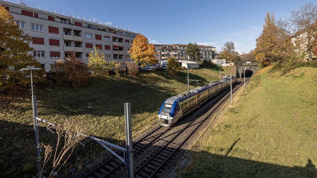 Ein blauer TER-Zug fährt auf Gleisen, die sich durch eine grüne Landschaft schlängeln, und nähert sich dem Eingang des Kannenfeldtunnels in Basel; im Hintergrund sind mehrstöckige Gebäude und herbstlich gefärbte Bäume zu sehen. Die Szene wird von der warmen Sonne des Oktobers beleuchtet, die Schatten wirft und die Farben der Umgebung hervorhebt.