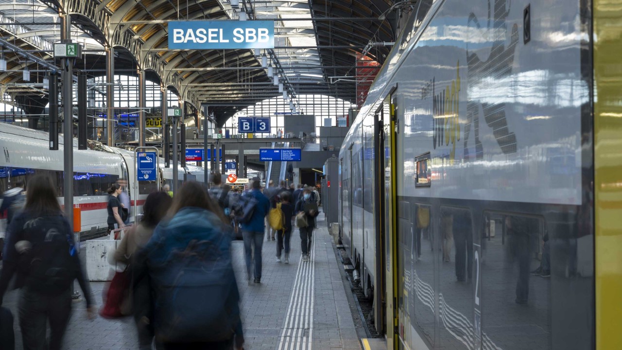 Der Bahnhof Basel SBB ist belebt mit Reisenden, die sich auf dem Perron bewegen. Zwei Züge stehen zu beiden Seiten des Bahnsteigs, während das komplexe Dach mit Metallträgern und offenen Bereichen natürliches Licht hereinlässt. Überkopfschilder zeigen Informationen zu den Perrons und Zielen.