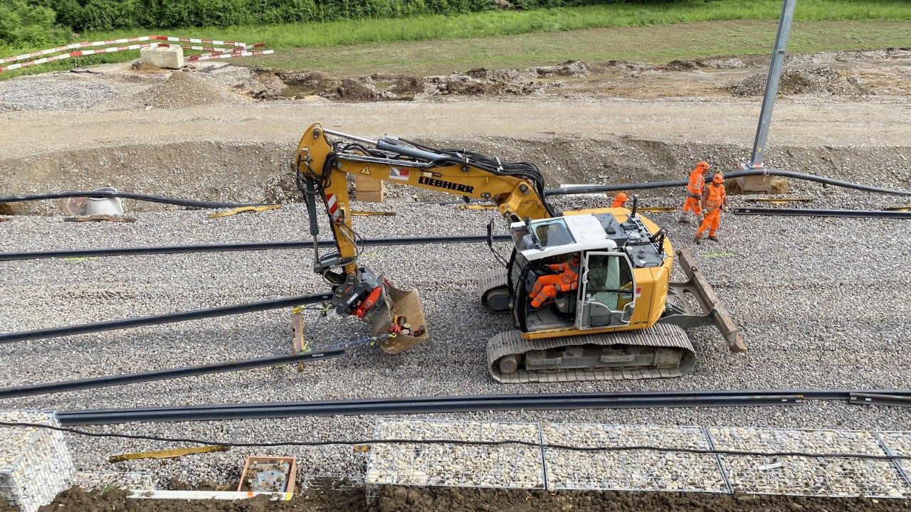 Ein gelber Bagger mit der Aufschrift ‚Liebherr‘ arbeitet auf einer Baustelle mit Schotterboden und mehreren Schienen. Zwei Personen in orangefarbener Schutzkleidung stehen daneben. Im Hintergrund sind Erdarbeiten und grüne Vegetation sichtbar.