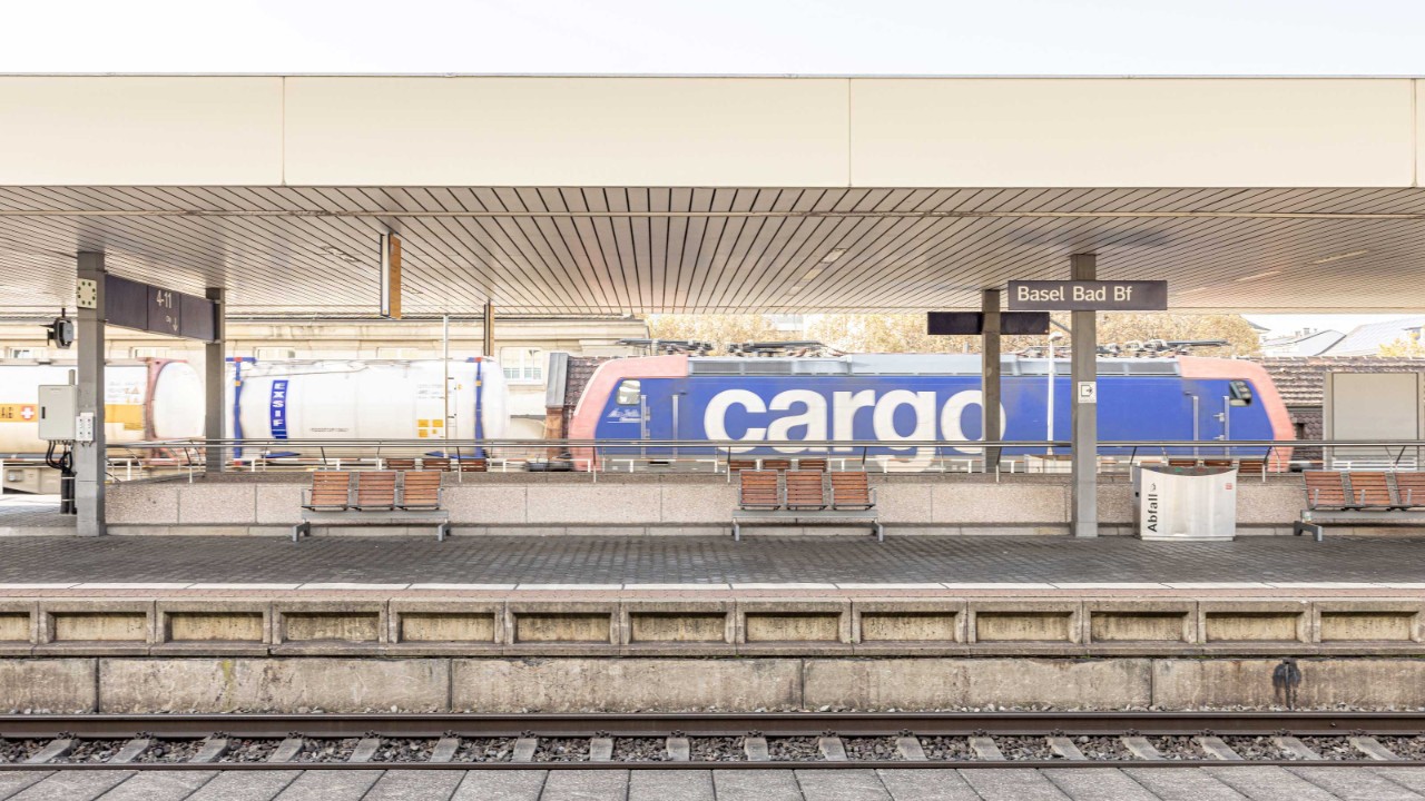 Am Basel Badischer Bahnhof steht ein blau-weisser Güterzug mit der Aufschrift “cargo”. Die Perrons sind leer, was auf wenig Betrieb hindeutet. Über dem Perron spannt sich ein grosses Dach.