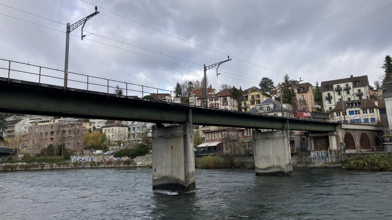 Das Bild zeigt die Reussbrücke Geissmatt in Luzern. Es ist eine Brücke mit Betonpfeilern, die über die Reuss führt. Im Hintergrund sind mehrstöckige Gebäude sichtbar. Die Brücke ist mit Oberleitungen ausgestattet, damit die Züge verkehren können.