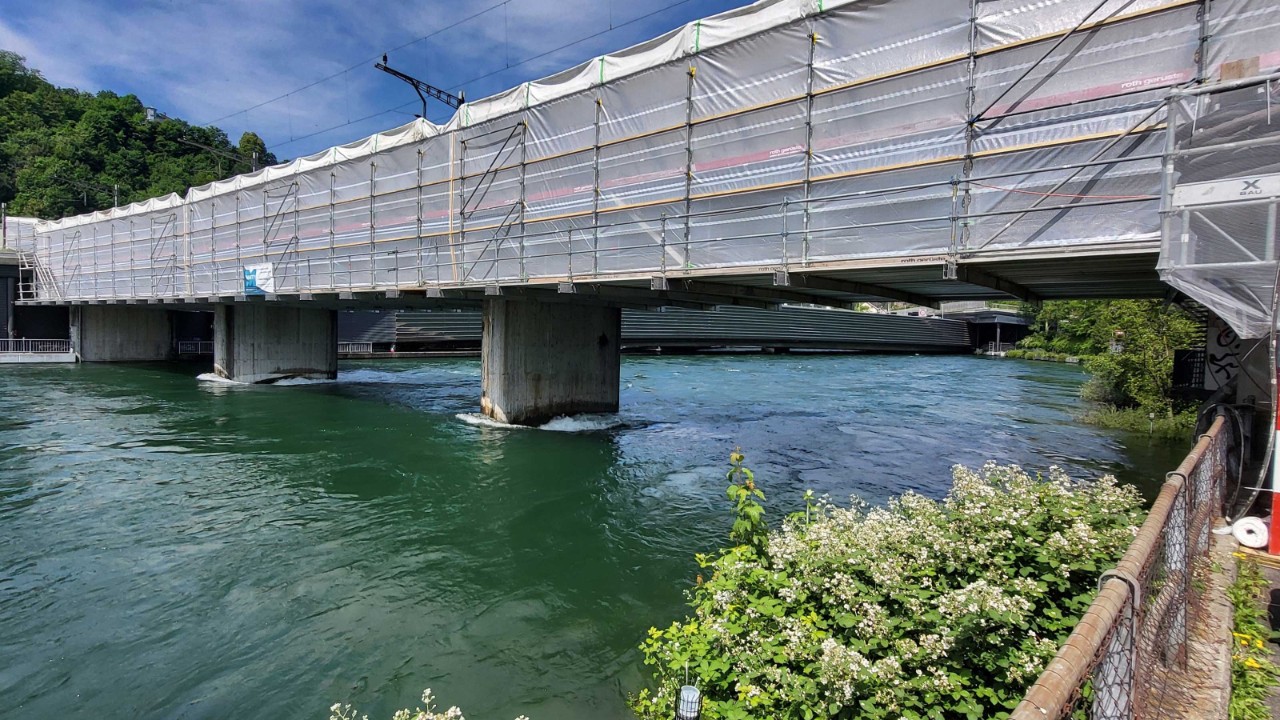 Das Bild zeigt die Geissmattbrücke in Luzern, wie sin in eingerüstetem Zustand die Reuss überspannt. Das Foto ist von der rechten Uferseite flussaufwärts aufgenommen. Vom eigentlichen Brückenbauwerk sind nur die Betonstützen zu sehen, der Brückenkörper ist vollständig eingerüstet. Das Gerüst ist mit einer weissen Folie umhüllt. Auffallend ist der hohe Wasserstand der Reuss: Das Foto ist Anfang Juni aufgenommen, zu einem Zeitpunkt da viele Gewässer in der Schweiz hohe Wasserstände aufweisen.