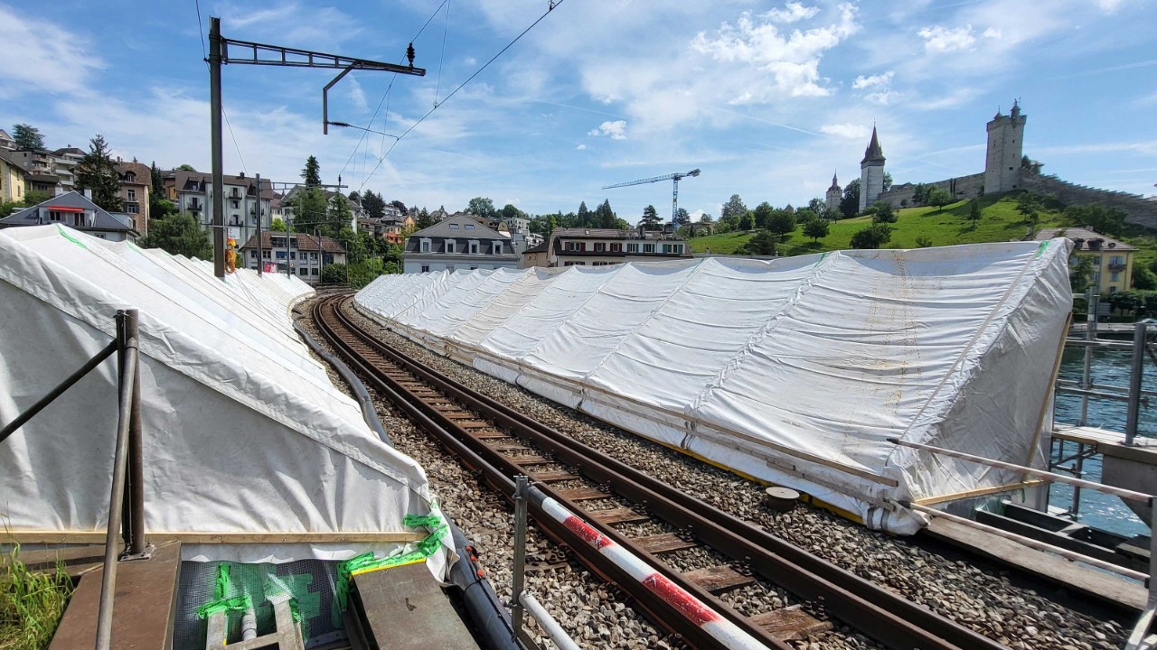 Das Bild zeigt die Oberseite der Geissmattbrücke in Luzern während der Bauarbeiten. Links und rechts der Fahrbahn ist das Brückenbauwerk eingerüstet und mit weisser Folie eingehaust. Das Foto ist von der linken Uferseite der Reuss aufgenommen. Im Hintergrund sind die Museggtürme und die Stadtmauer Luzerns zu sehen.