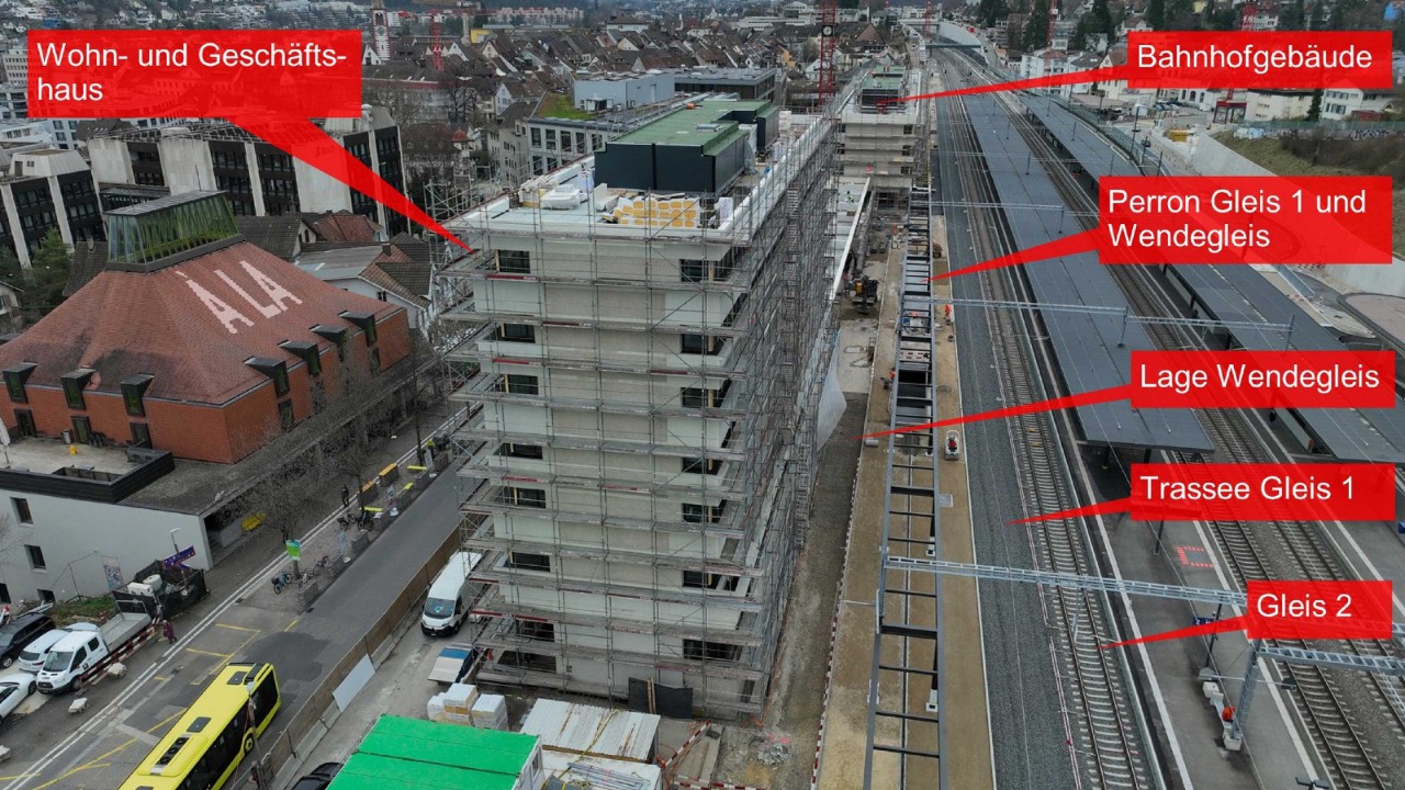 Das Luftbild zeigt den Bahnhof Liestal mit Blick in Richtung Olten. Im Vordergrund ist ein mehrstöckiges Gebäude im Bau zu sehen, während im Hintergrund das Bahnhofsgebäude sichtbar ist. Auf dem Trassee des Gleis 1 liegt erst der Schotter. Das Perron ist im Bau, vom Perrondach steht das Metallgerüst.