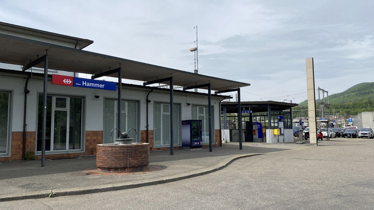 Das Foto zeigt das Bahnhofsgebäude des Bahnhofs Olten Hammer mit überdachtem Vorplatz. Rechts daneben steht ein pavillonartiger Bau, in dem sich die Spiralrampe zur Bahnhofsunterführung befindet. Die Aufnahme zeigt den heutigen Zustand des Zugangs im bestehenden Umfeld.