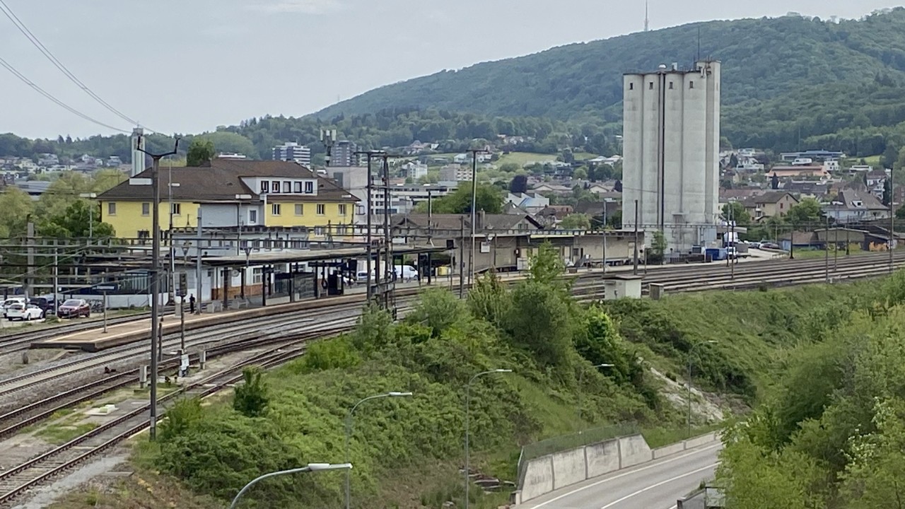 Das Foto zeigt den Bahnhof Olten Hammer mit mehreren Gleisen, Perrons und Oberleitungen aus leicht erhöhter Perspektive. Rechts sind markante Silogebäude und weitere Betriebsbauten zu sehen, im Hintergrund Wohnquartiere und bewaldete Hügel. Die Aufnahme verortet die Bahnanlagen im bestehenden städtischen und landschaftlichen Umfeld.