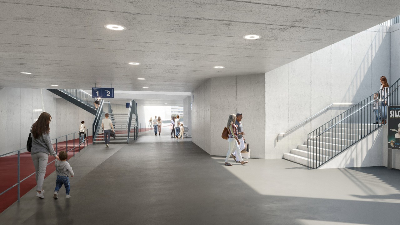 Die Visualisierung zeigt eine breite, helle Unterführung mit Betonflächen, Deckenleuchten und klar geführten Gehwegen. Sichtbar sind Treppenanlagen sowie Personen, die sich durch den Raum bewegen. Geländer, Wegmarkierungen und offene Blickbeziehungen unterstützen die Orientierung für Reisende.
