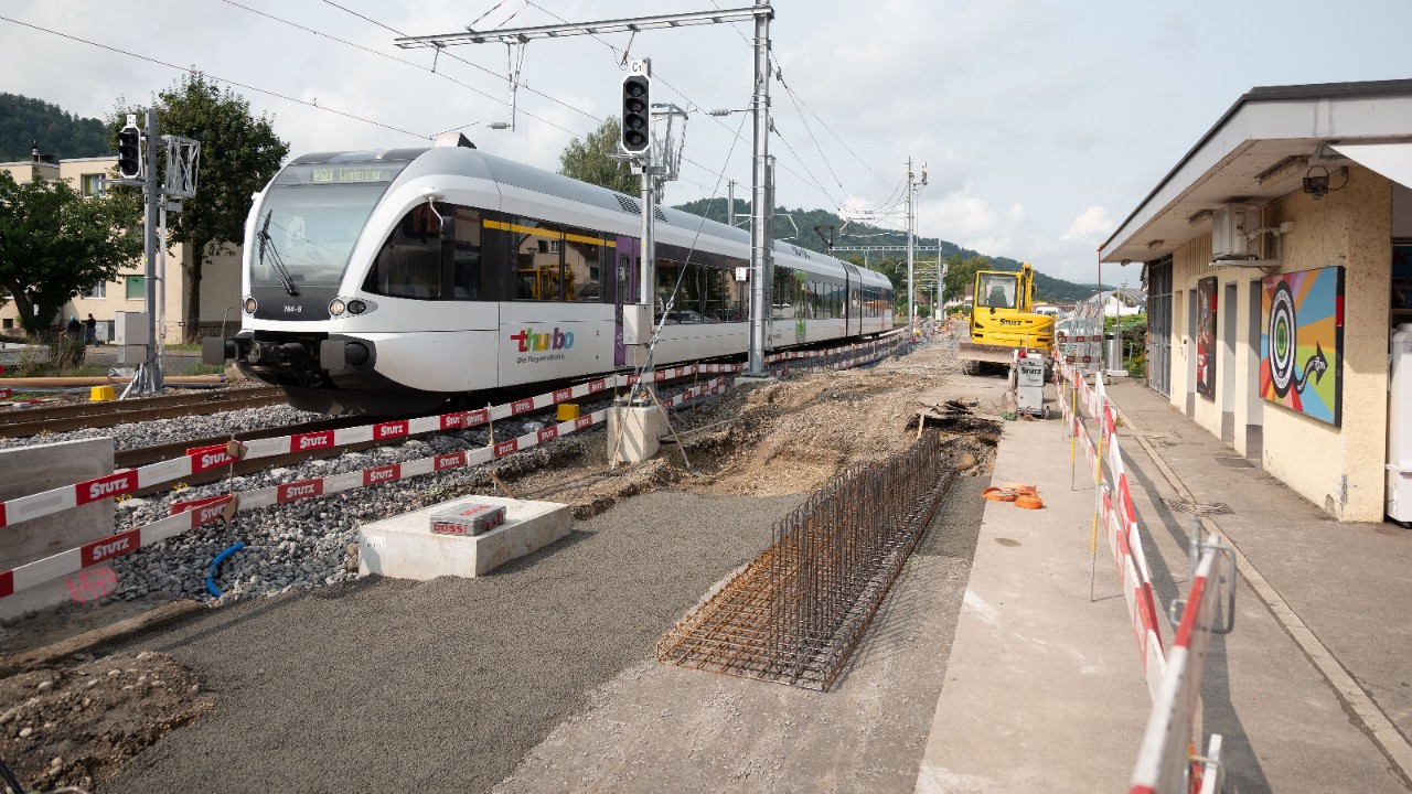 Das Foto zeigt einen Thurbo-Zug während der Bauzeit auf dem Bahnhof Pfungen. Das Perron ist teilweise aufgerissen, ein Bagger hebt einen Schacht aus.