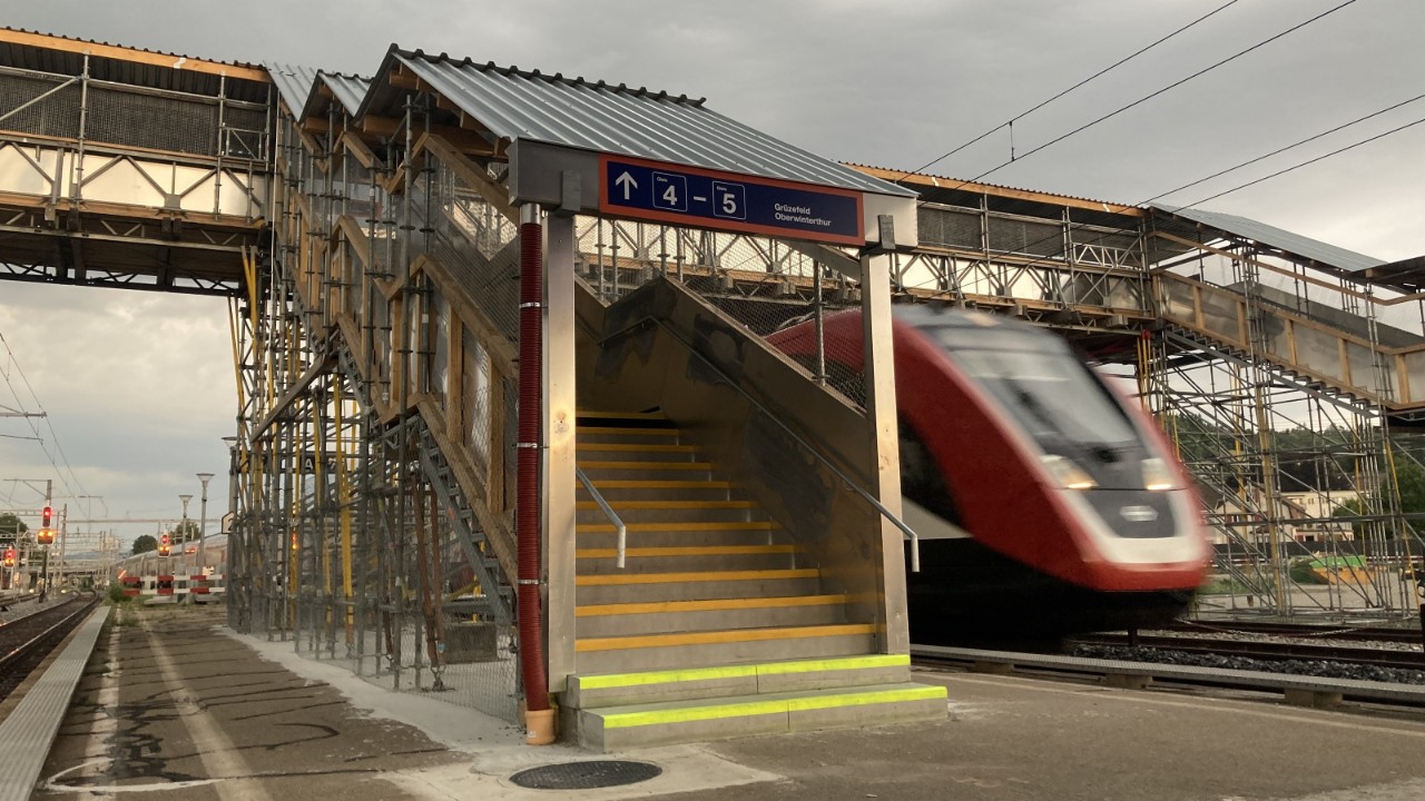 Das Bild zeigt die provisorische Passerelle am Bahnhof Winterthur Grüze, die aus Baugerüst-Elementen gefertigt ist. Sowohl Treppen als auch die Gleisquerung sind überdacht. Die provisorische Passerelle hat keine Rampen und ist nicht barrierefrei.