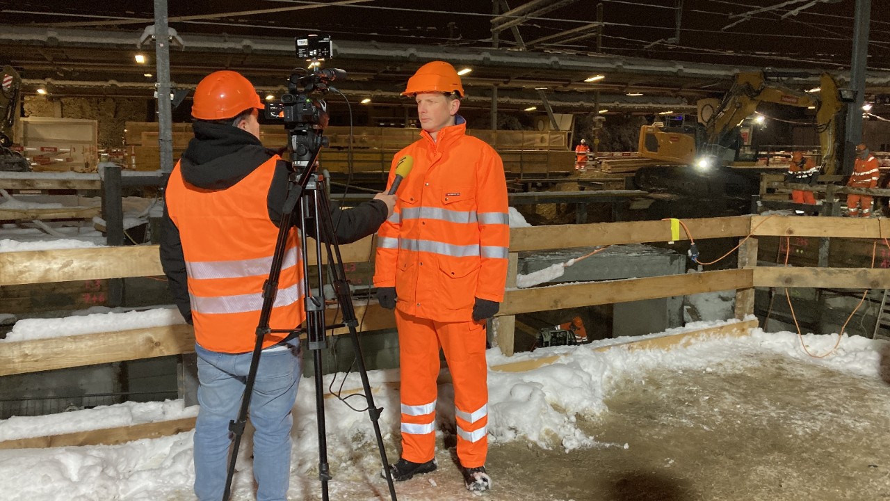 Das Foto zeigt Manuel Meier der vor der Baustellenabschrankung ein Fernsehinterview gibt. Im Hintergrund sind Bauarbeiter und ein Bagger im Einsatz und die Perrondächer des Bahnhofs Grüze sind zu sehen.
