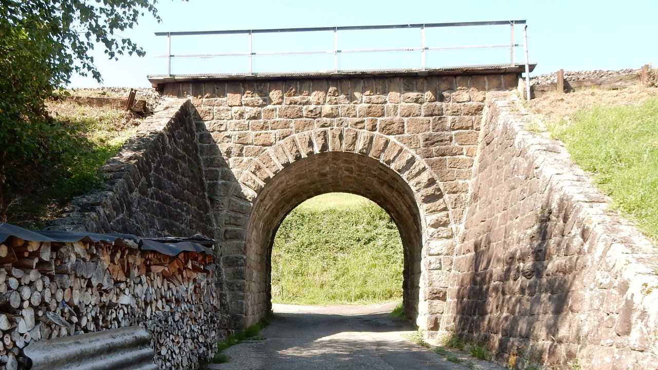 Das Foto zeigt den Natursteinbogen der Wegunterführung sowie die Flügelmauern. Über den Bogen, der von der Bauweise her an einen Rundbogen aus dem Mittelalter erinnert, verläuft die Bahnstrecke.