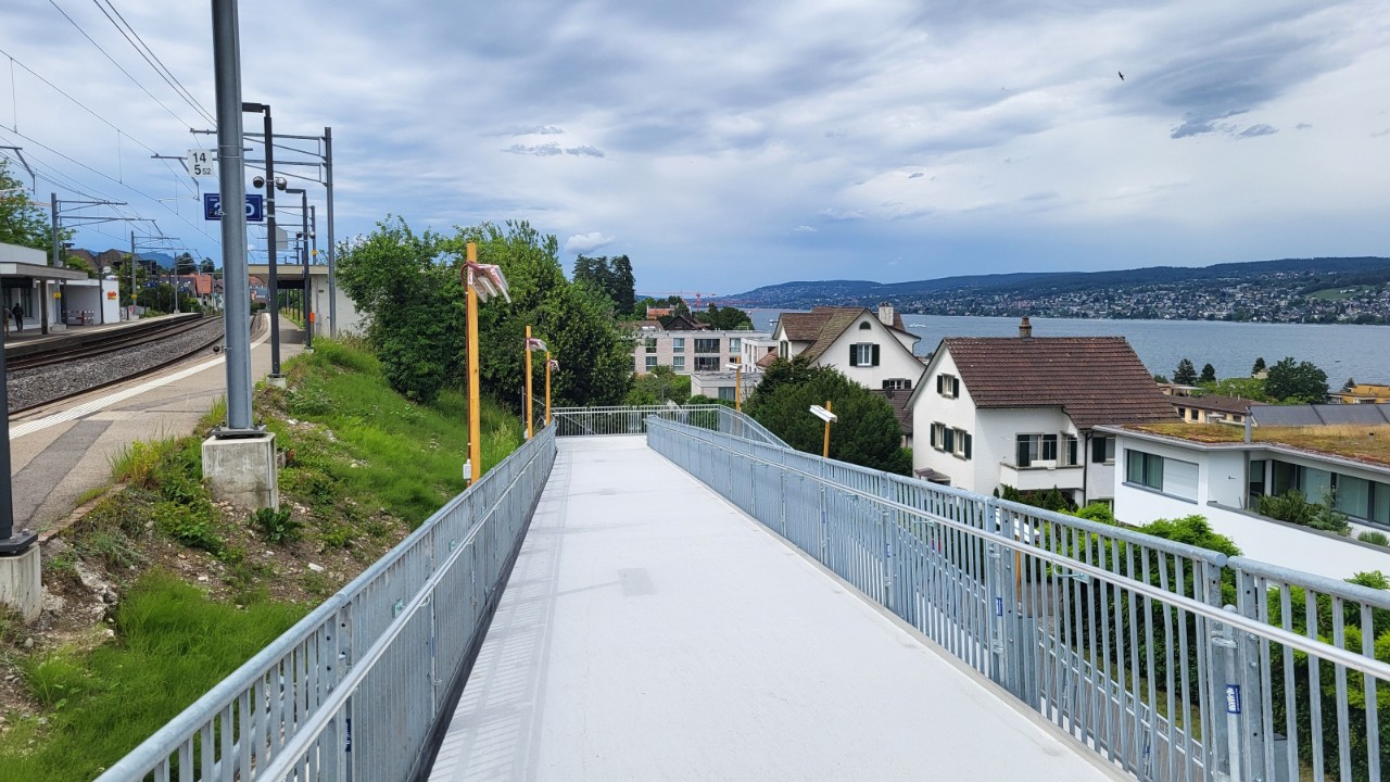 Das Foto zeigt die provisorische Rampe am Bahnhof Oberrieden Dorf, Blickrichtung vom oberen Teil der Rampe nach unten. Der weisse Belag der breiten Rampe steht im Zentrum des Bildes, links und rechts sind Geländer mit Handläufen zu sehen. Am linken bildrand ist das Perron Gleis 2 zu sehen, an welches die Rampe anschliesst. Im Hintergrund liegt der malerische Zürichsee.