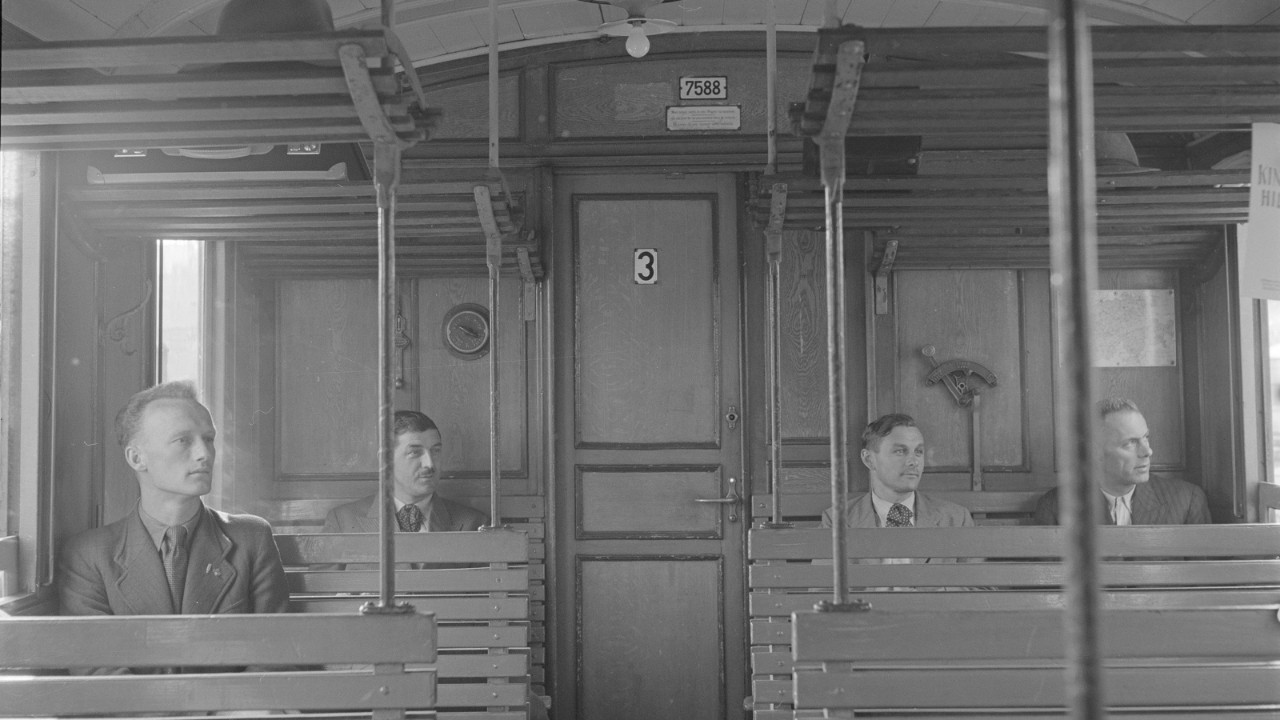 Interior of a third-class carriage with wooden benches, hat racks and passengers.
