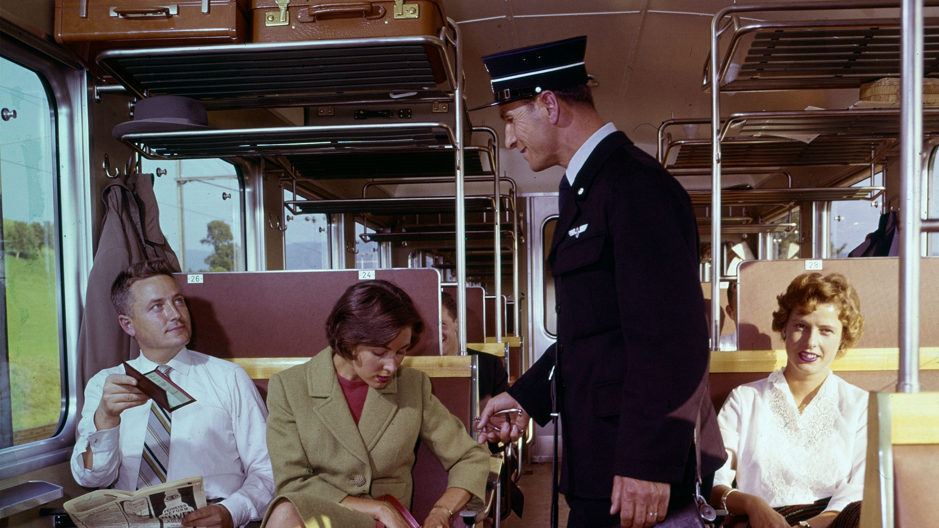 A member of train crew wearing a hat and carrying a bag inspects passengers’ tickets on a train, 1959.