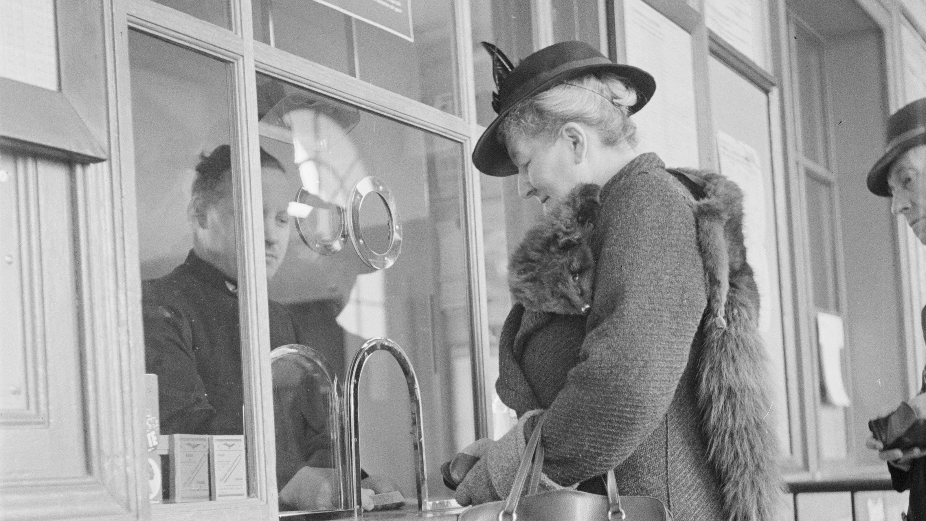 A customer asks for advice at a ticket counter. A glass panel with an open hatch separates the customer from the employee, 1938.