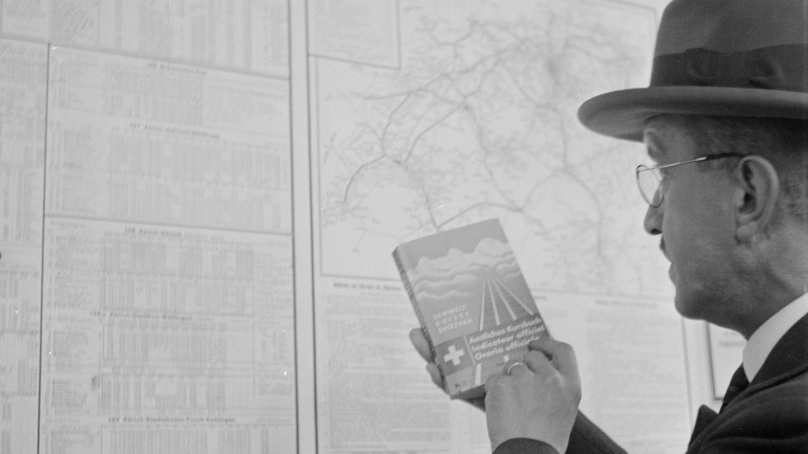 Black and white photograph of a passenger in 1936 holding an official timetable and standing in front of a printed timetable.