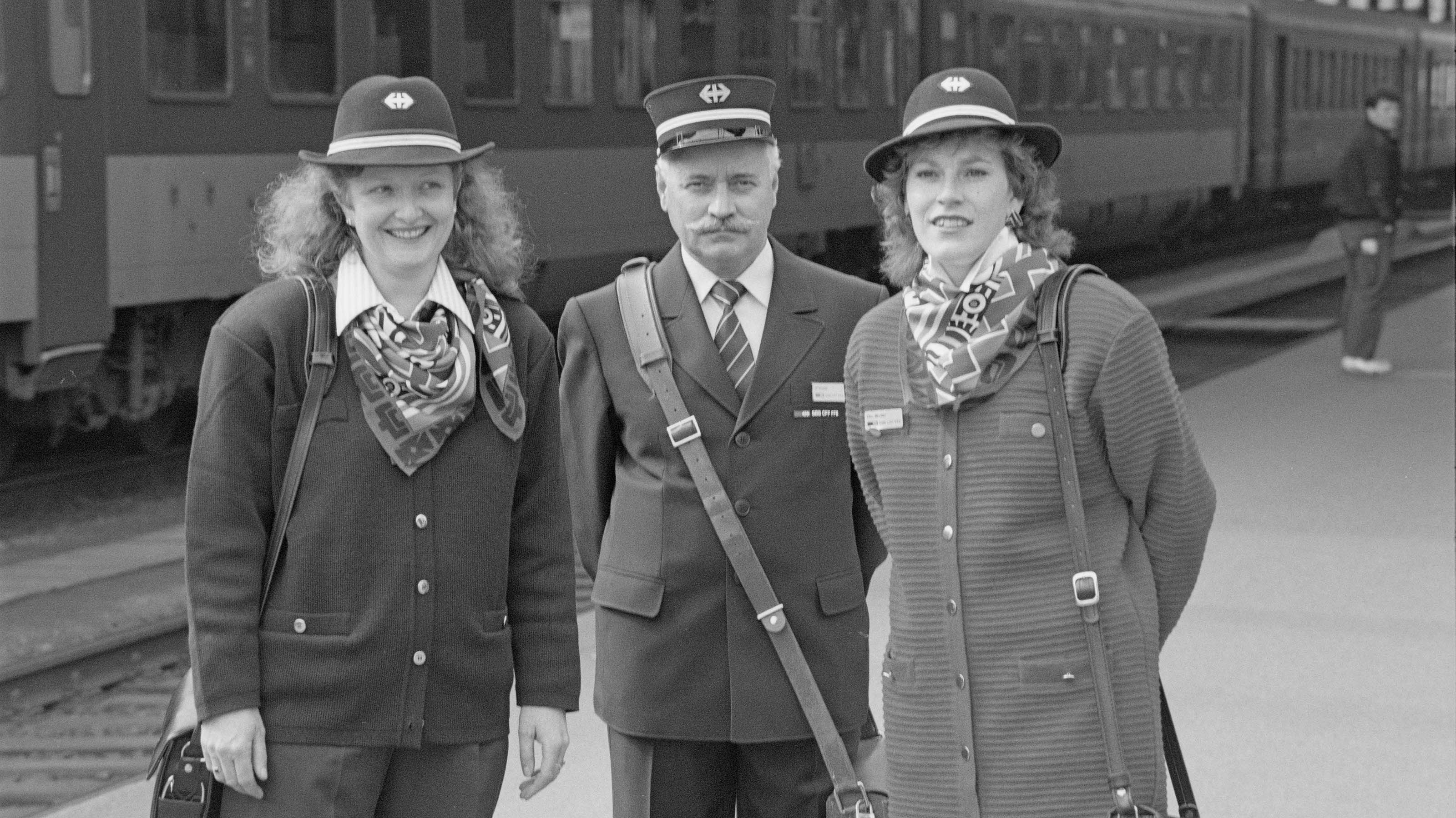 Black and white photograph of three uniformed passenger attendants standing on a platform, 1991.