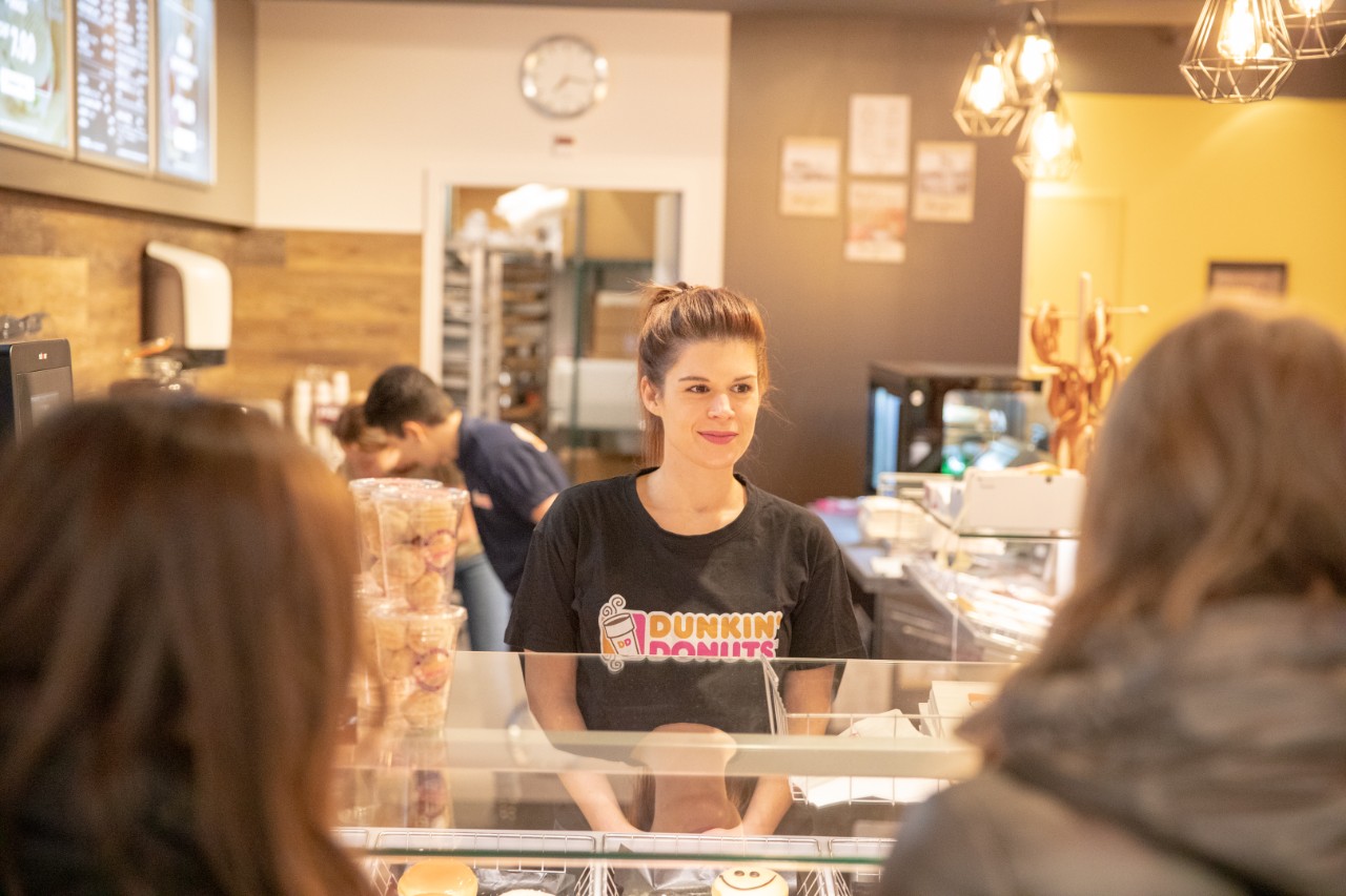 Eine Verkäuferin von Dunkin’ Donuts bedient lächelnd zwei Kundinnen.
