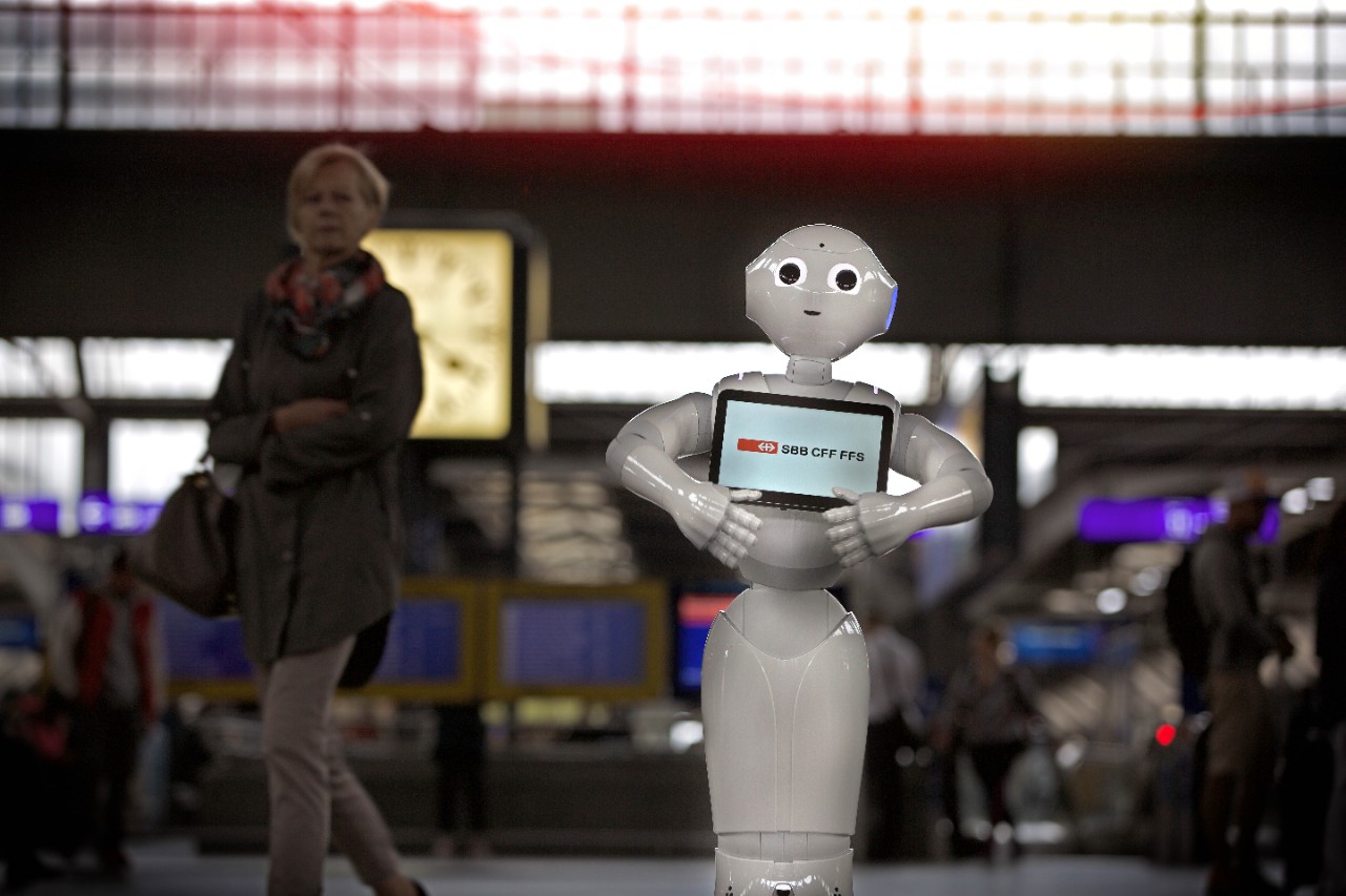 Roboter „Pepper“ steht mit einem „SBB CFF FFS“ auf der Brust im Zürich HB. Im Hintergrund geht eine Kundin vorbei.