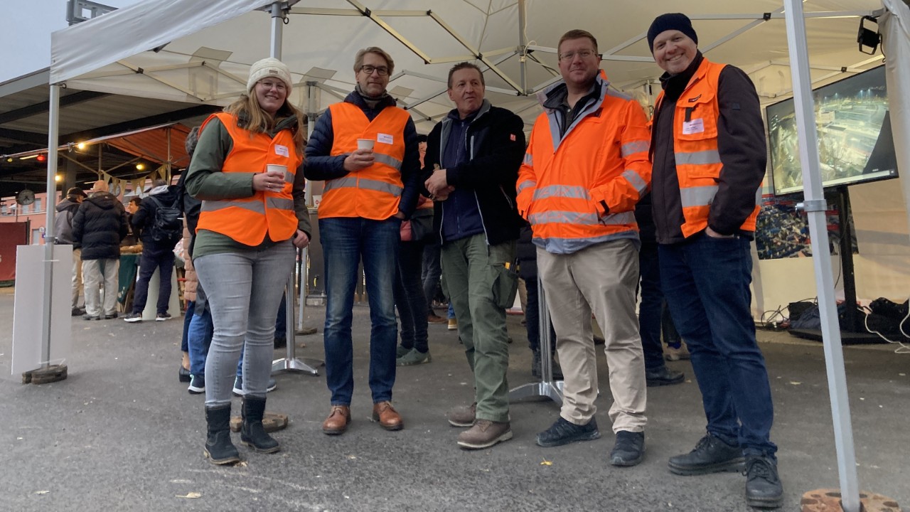 Das Foto zeigt fünf Mitglieder des Bauprojekt-Teams in oranger Sicherheitskleidung unter dem Dach des Infozelts, das fürs Bahnhoffest auf dem Bahnhofplatz Muttenz aufgebaut wurde.